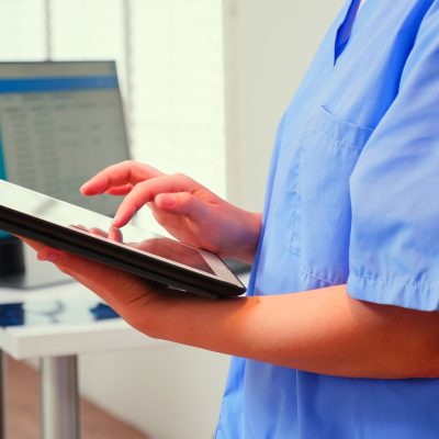 Close up of assistant holding tablet and analysing teeth digital radiography of patient standing in stomatologic clinic, while doctor is working in background. Nurse using modern technology